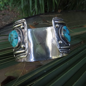 May include: A silver cuff bracelet with two turquoise stones set in silver. The bracelet is decorated with intricate silver designs.