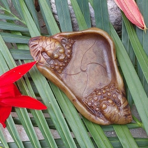 May include: A brown, carved stone dish with a design of grapes and a stylized insect. The dish is resting on a bed of green palm leaves.