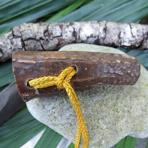 May include: A handcrafted wooden toggle with a textured, bark-like appearance. A bright yellow cord is threaded through a hole in the toggle and tied. The toggle rests on a light green stone, with a blurred background of greenery and a piece of wood.