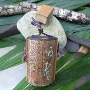 May include: A small, cylindrical wooden box with a dark brown finish and a purple cord. The box has Japanese characters written on it and is sitting on a bed of green leaves.