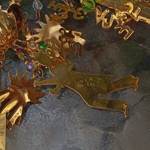 May include: Gold-colored metal ornament with a whimsical design featuring a figure with outstretched arms and legs. The ornament is decorated with beads and crystals in shades of green, purple, and clear. The ornament is signed "Lunch At The Ritz" on the back.