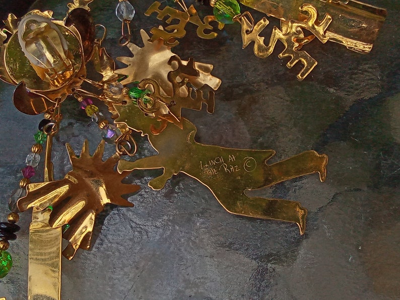 May include: Gold-colored metal ornament with a whimsical design featuring a figure with outstretched arms and legs. The ornament is decorated with beads and crystals in shades of green, purple, and clear. The ornament is signed "Lunch At The Ritz" on the back.