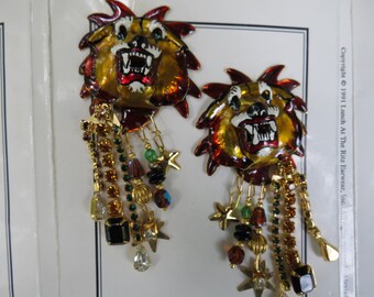 May include: A pair of vintage earrings featuring a lion's head design with a fierce expression. The earrings are gold, red, and white with dangling strands of colorful beads, crystals, and star charms. Copyright 1991 Lunch At The Ritz Earwear, Inc.