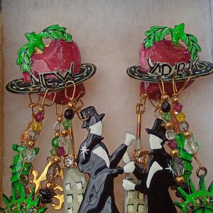 May include: A pair of dangle earrings featuring a red apple with green leaves and the words "New York" and "York" on the front. The earrings are adorned with gold-colored chains, green and red beads, and black and white figures.