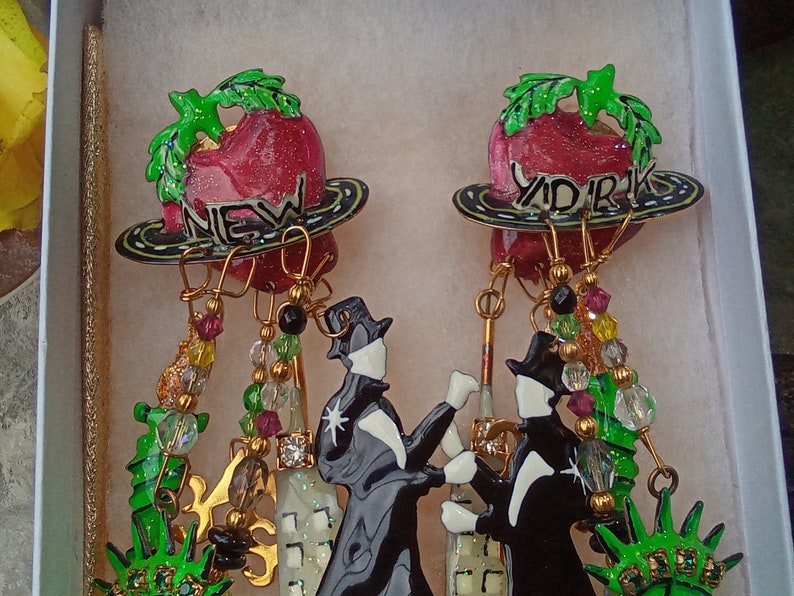 May include: A pair of dangle earrings featuring a red apple with green leaves and the words "New York" and "York" on the front. The earrings are adorned with gold-colored chains, green and red beads, and black and white figures.