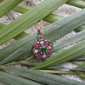 May include: A silver pendant with a green and red gemstone flower design. The pendant is on a green leafy background.
