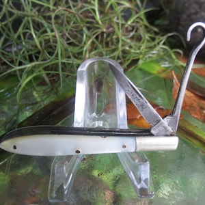 May include: A vintage pocket knife with a mother-of-pearl handle and a silver-colored blade. The knife has a hook-shaped tool and is displayed on a clear plastic stand. The blade is partially open, revealing the sharp edge.
