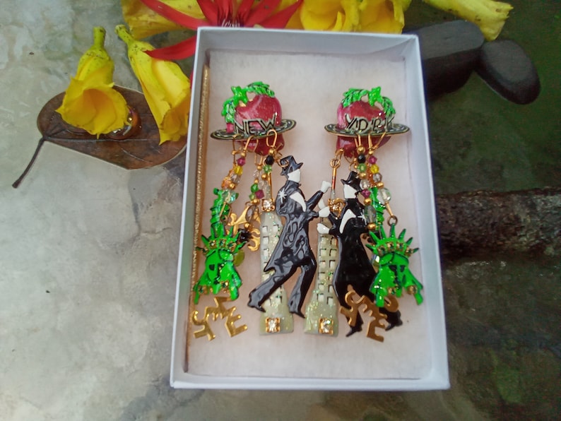 May include: A pair of dangle earrings with a red apple charm with the words "New York" on it. The earrings also feature a black and white silhouette of a couple dancing, a green Statue of Liberty charm, and gold accents.