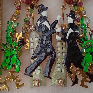 May include: A pair of earrings featuring a black and white enamel figure of a person dancing, a green enamel figure of the Statue of Liberty, and a white and gold enamel building with a sparkly finish. The earrings are attached to a gold chain with green, red, and gold beads.