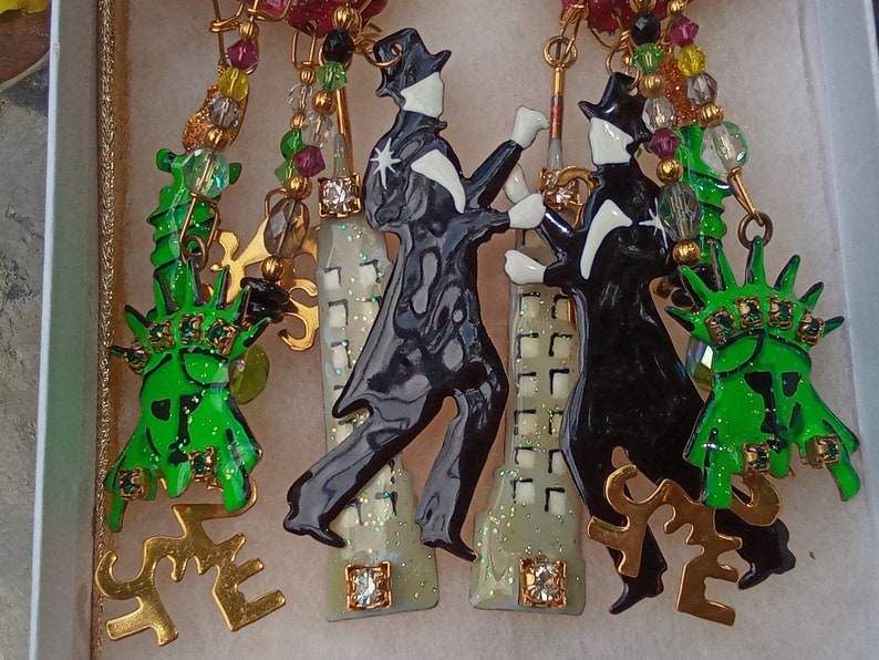 May include: A pair of earrings featuring a black and white enamel figure of a person dancing, a green enamel figure of the Statue of Liberty, and a white and gold enamel building with a sparkly finish. The earrings are attached to a gold chain with green, red, and gold beads.