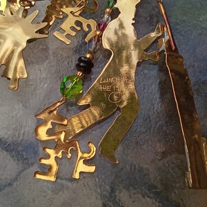 May include: Gold-colored metal ornament with a silhouette of a person dancing, with the words "Lunch at the 13th" inscribed on the ornament. The ornament is decorated with green and black beads.