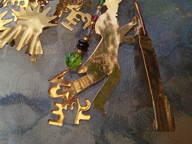 May include: Gold-colored metal ornament with a silhouette of a person dancing, with the words "Lunch at the 13th" inscribed on the ornament. The ornament is decorated with green and black beads.