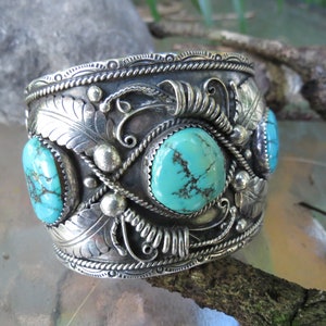 May include: A silver cuff bracelet with intricate designs and turquoise stones. The bracelet is decorated with a floral pattern and has a rope-like border.