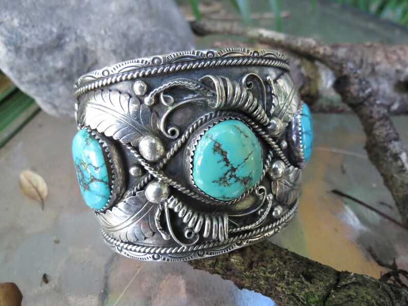May include: A silver cuff bracelet with intricate designs and turquoise stones. The bracelet is decorated with a floral pattern and has a rope-like border.