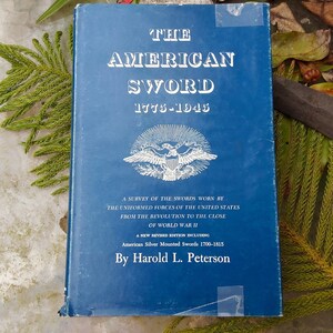 May include: A blue hardcover book titled "The American Sword 1775-1945" with a white image of an eagle on the front cover. The book is about a survey of the swords worn by the uniformed forces of the United States from the Revolution to the close of World War II.