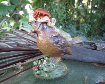 May include: A decorative bird figurine with a brown body, wearing a brown hat with lace trim and a brown harness. The bird stands on a green base with ribbon and rose accents. A charming, vintage-style ornament.