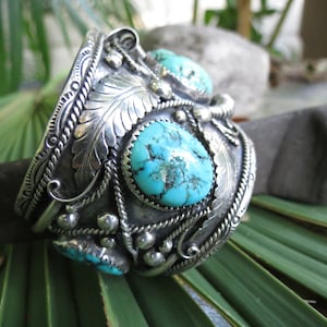 May include: A silver cuff bracelet with turquoise stones set in a detailed design. The bracelet features intricate silverwork with a floral motif and a rope-like border.