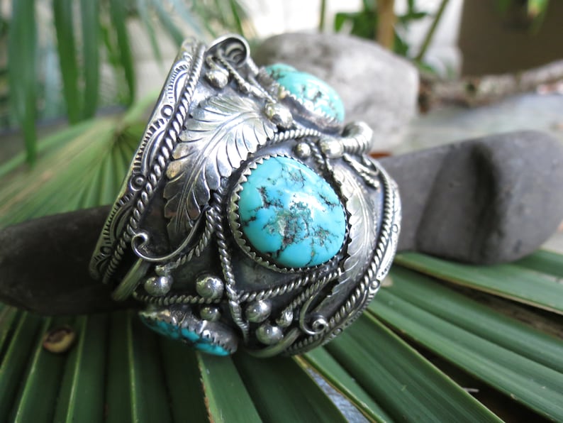 May include: A silver cuff bracelet with turquoise stones set in a detailed design. The bracelet features intricate silverwork with a floral motif and a rope-like border.