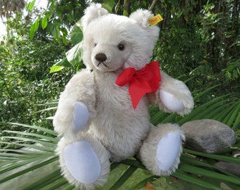 May include: A white plush teddy bear with a red bow sitting on a green palm leaf. The bear has a button nose and white paw pads.