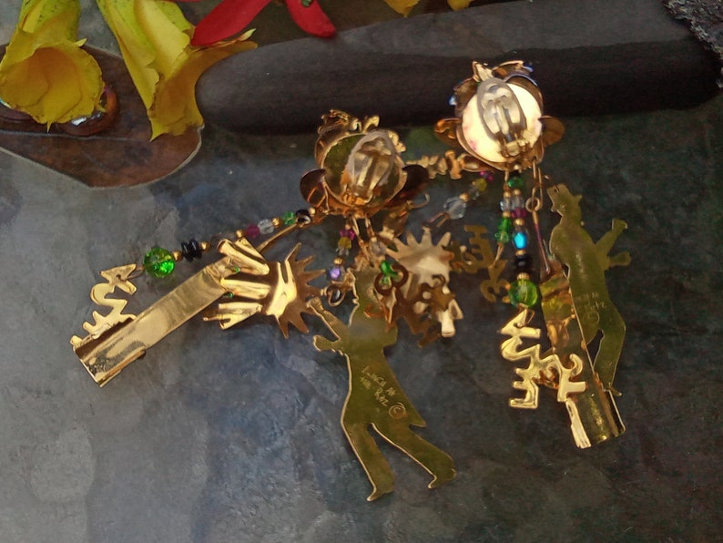 May include: A pair of gold-tone dangle earrings with a variety of charms, including a silhouette of a person, a palm tree, and a star. The earrings are adorned with colorful beads and crystals.