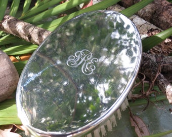 May include: Oval silver compact mirror with an engraved monogram. The mirror reflects the surrounding foliage and sky. The compact has a polished, reflective surface and a decorative edge. The monogram appears to be the letters "R" and "K".