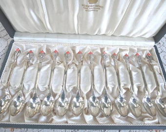 May include: A set of 12 silver teaspoons with coral-colored handles in a velvet-lined box. The box is labeled "A.B. Nordiska Kompaniet,  H.K.H. Hovleverantör, Stockholm."