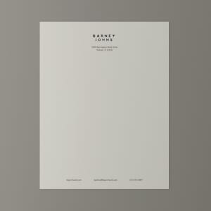Könnte beinhalten: Ein cremefarbenes Briefpapier mit dem Text "BARNEY JOHNS" in einer minimalistischen schwarzen Schriftart oben. Darunter befinden sich Adresse und Kontaktdaten. Das Papier ist auf einem grauen Hintergrund zentriert.
