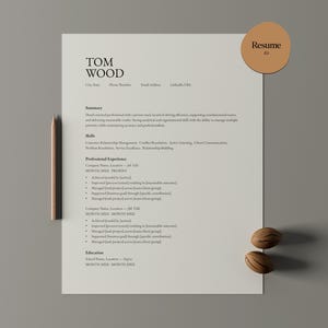 Puede incluir: Una plantilla de currículum vitae con el nombre "TOM WOOD" en la parte superior. El documento incluye secciones para resumen, habilidades, experiencia profesional y educación. Un lápiz de madera y dos nueces decorativas se colocan cerca del documento. Una pegatina circular con la palabra "Resume" está en la parte superior derecha.