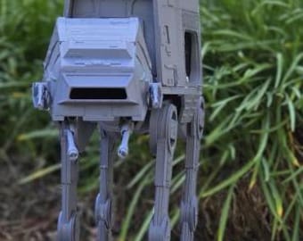 Peut inclure: Modèle gris d'un AT-AT de Star Wars, posé sur une surface en pierre avec un feuillage vert en arrière-plan. Le modèle présente des détails, notamment un corps rectangulaire, un avant incliné et quatre pattes articulées.