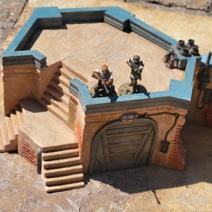 May include: A miniature diorama of a building with a tan exterior, blue trim, and a flat roof. Two small figures stand on the roof, holding weapons. The building has stairs, a door, and various architectural details, creating a detailed, realistic scene.