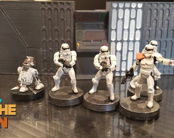 May include: A group of five white Star Wars Stormtrooper figurines, including one with an orange shoulder pad, standing on black bases. A small white droid figurine is also included. The figurines are set against a backdrop of a gray and white wall with the text "Imperial Interior Tosche Station www.ttterrain.com".