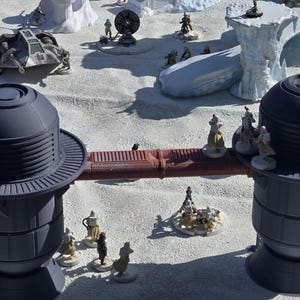 May include: A tabletop diorama depicting a snowy landscape with miniature structures and figures. The scene includes a vehicle, buildings, and various figurines, all set against a white backdrop, creating a miniature battle scene.