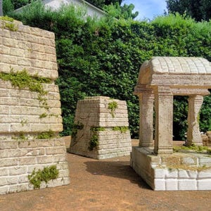 Può includere: Tre strutture in pietra in miniatura, che ricordano rovine antiche, con una texture a mattoni e accenti di muschio. Le strutture sono disposte su una superficie marrone.