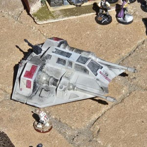 May include: A detailed model of a Star Wars Snowspeeder in shades of gray and white, with red accents. The model features intricate detailing, including guns and cockpit windows. A small figure of a droid is also present.