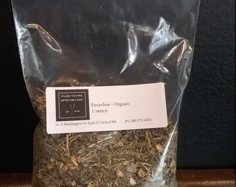 May include: A clear plastic bag filled with dried green herbs labeled "Feverfew - Organic 1 ounce" from Olde Thyme Apothecary. The label also includes the business address and phone number: 51 S Washington St Suite D Oxford MI (P) 248-572-6404.