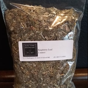 May include: A clear plastic bag filled with dried raspberry leaves. The bag has a label that reads "Raspberry Leaf 1 ounce" and includes the business name "Olde Thyme Apothecary" and contact information.