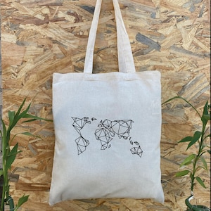 May include: A cream-colored tote bag with a black geometric world map design. The bag has long, matching handles and is hanging against a wood-grain background. The bag is made of a sturdy fabric.