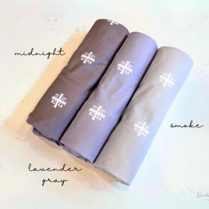 Pu&ograve; includere: Tre campioni di tessuto in tonalit&agrave; di grigio, etichettati "midnight", "lavender gray" e "smoke". Ogni campione presenta una croce bianca ricamata.