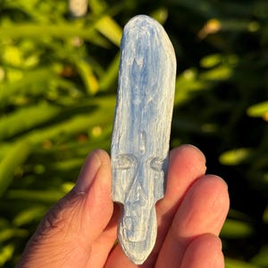 May include: A carved blue celestite crystal in the shape of a face. The crystal has a smooth, polished surface and is held in a hand.