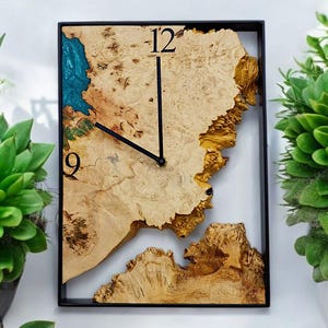 Può includere: Orologio da parete quadrato in legno con un design a venature naturali e lancette nere. Il quadrante presenta una forma unica con accenti di resina blu e i numeri 12 e 9 visibili. L'orologio è incorniciato in un quadrato nero.