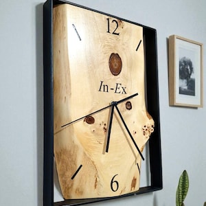 Può includere: Un orologio quadrato in legno con un quadrante in legno chiaro e naturale e lancette e numeri neri. Il quadrante dell'orologio ha il testo "In-Ex" ed è racchiuso in una cornice di metallo nero. L'orologio è montato su una parete grigio chiaro.