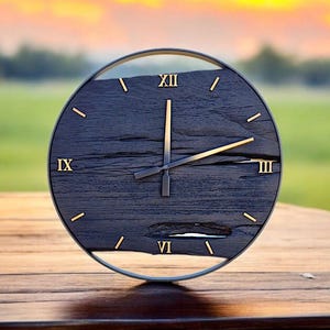 May include: A round, black wooden clock with gold-colored Roman numerals and hands. The clock face has a distressed wood grain appearance and is set in a thin, silver-colored frame. The clock is displayed on a wooden surface.