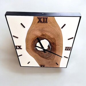 May include: A square wall clock with a black frame and a white face. The clock face features a natural wood grain design with Roman numerals and black clock hands. The wood grain has a unique, organic shape.