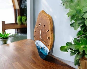 May include: A wooden wall clock with a natural wood grain finish. The clock face features gold Roman numerals and hands. The bottom portion is a blue and white resin design, resembling ocean waves. The clock measures approximately 12 inches in diameter.