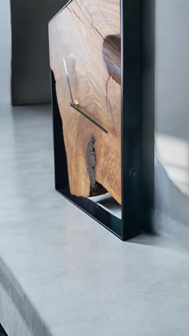 May include: A square wooden clock with a black metal frame. The clock face is made of natural wood with a unique grain pattern. The clock hands are black and gold.