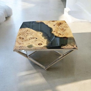 Op de afbeelding: Vierkante salontafel met een uniek ontwerp. Het tafelblad heeft een licht houtnerfpatroon met donkerblauwe harsaccenten. De tafel heeft een gepolijste metalen basis met een geometrisch ontwerp.