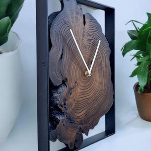 May include: A unique wall clock featuring a dark brown, cross-section of a tree trunk with visible growth rings. The clock face is set within a black rectangular frame. White clock hands indicate the time. A decorative piece for home decor.