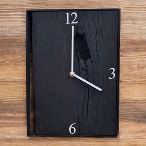 May include: A square, black wooden wall clock with white numerals at 12, 3, and 6. The clock hands are white. The clock is set against a wooden background. The clock measures approximately 12 inches by 10 inches.