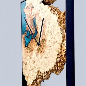 Könnte beinhalten: Quadratische Wanduhr mit einem Zifferblatt aus Naturholz und schwarzen Zeigern. Das Holz hat ein einzigartiges, strukturiertes Muster mit Bereichen aus blauem Harz. Die Uhr ist in einem schwarzen Metallrahmen eingefasst und schafft ein modernes, künstlerisches Design.