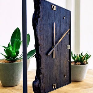 Può includere: Un orologio quadrato in legno scuro con numeri romani e lancette dorate. Il quadrante ha un bordo naturale su un lato ed è incorniciato in nero. L'orologio è esposto su un tavolo di legno con piante in vaso.